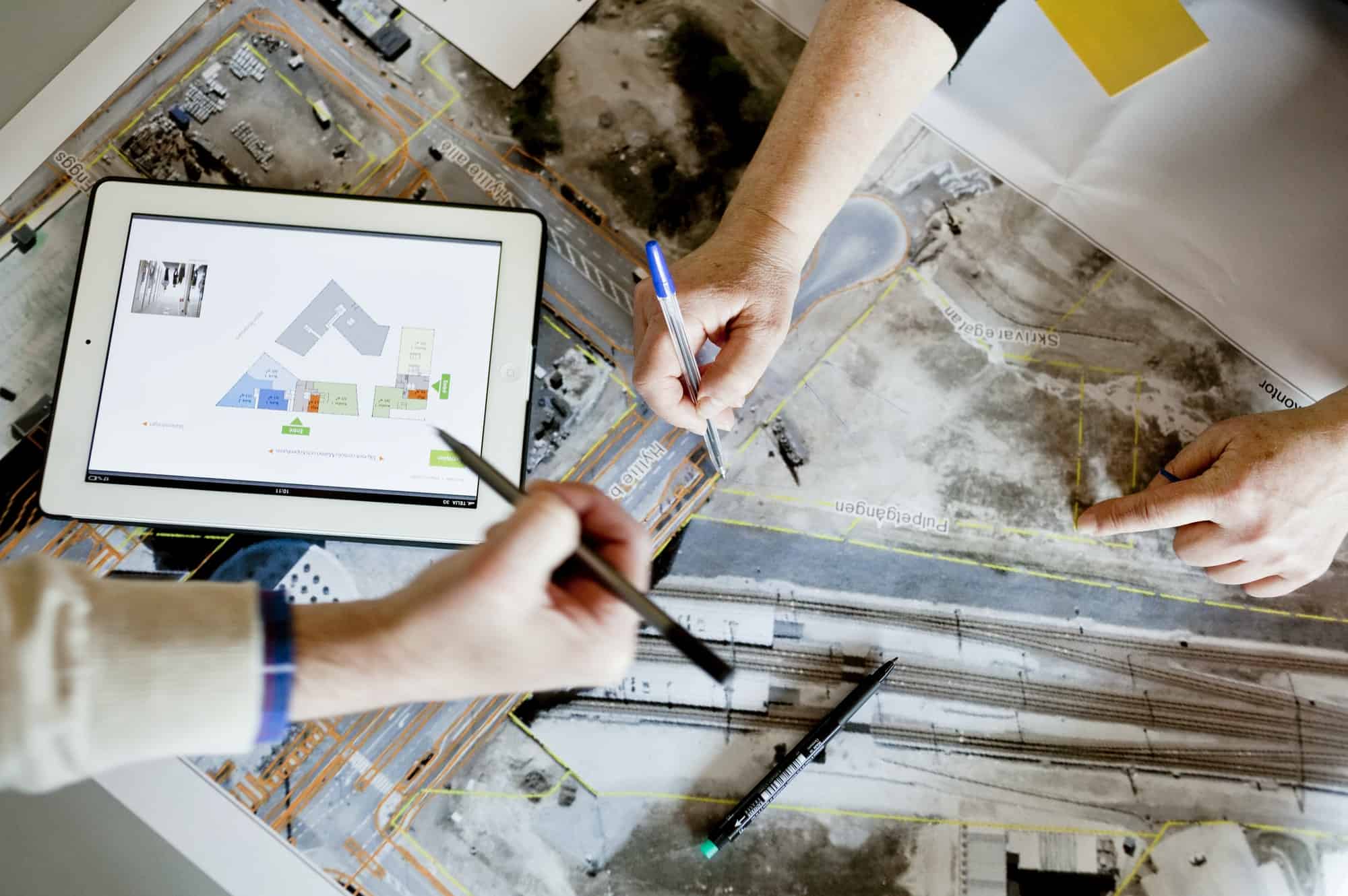 Two people, including a surveyor, collaborate over a map and architectural plans displayed on a tablet. One points with a pen while the other holds a marker, suggesting a planning or design discussion, with maps and plans spread across the table.