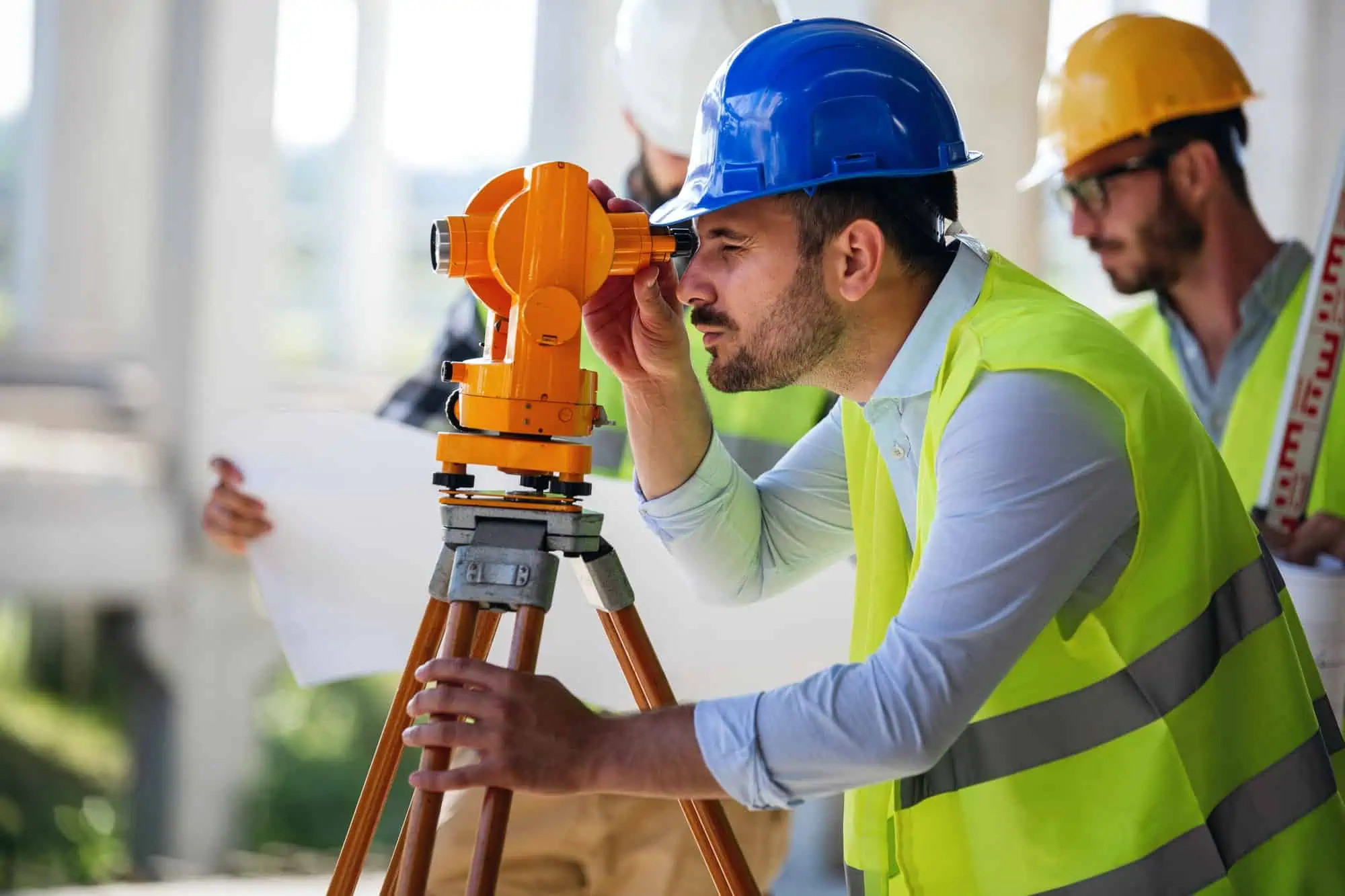 A construction worker in a blue hard hat and reflective vest uses a theodolite on a tripod, showcasing precision as a land surveyor. In the background, two other workers in hard hats and vests study construction plans at the building site.