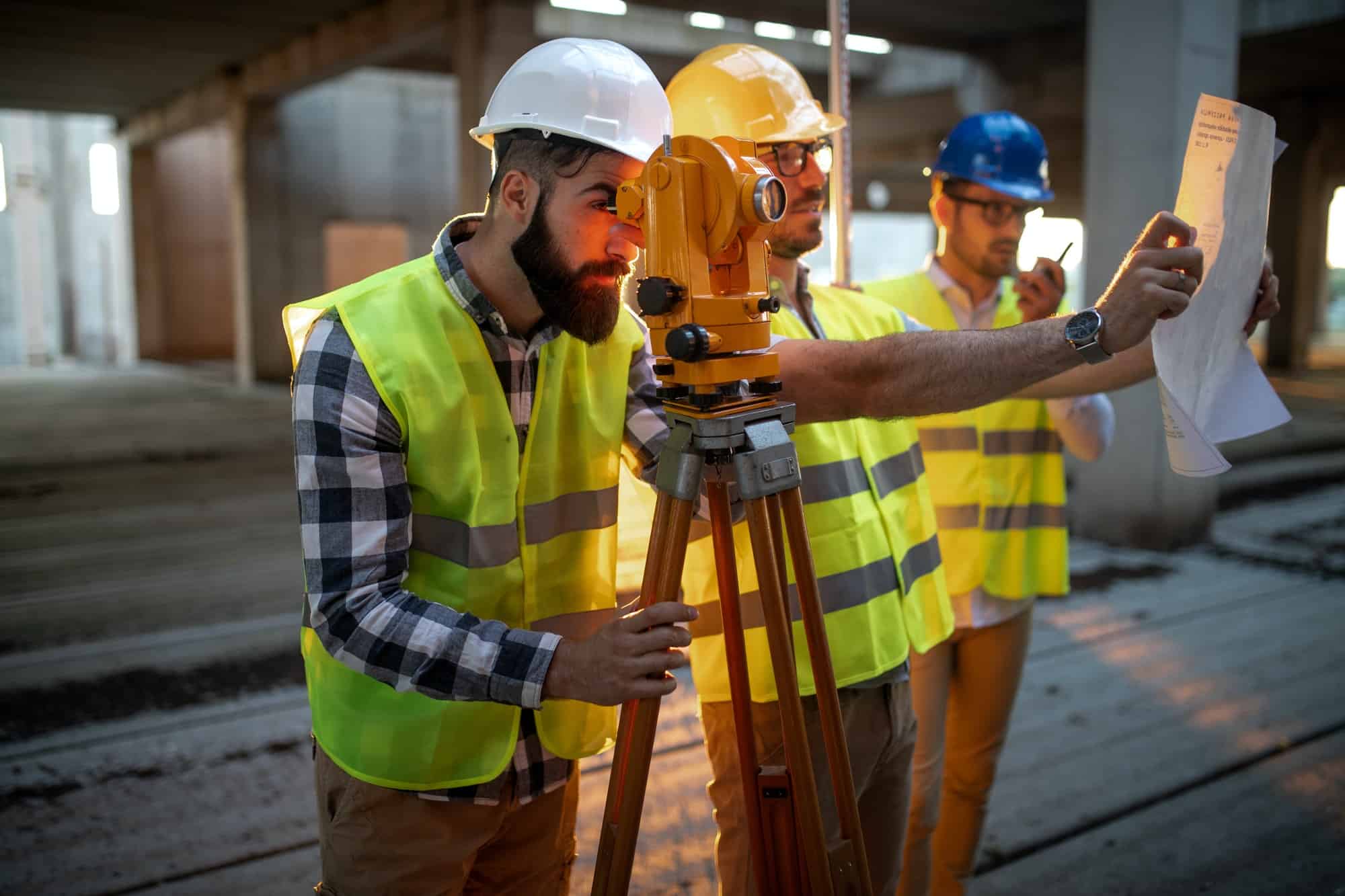 Three construction workers in hard hats and reflective vests use a theodolite as a land surveyor to survey a site. One holds a blueprint, while another looks through the theodolite. They are inside a partially built structure, ensuring precision in every measurement.