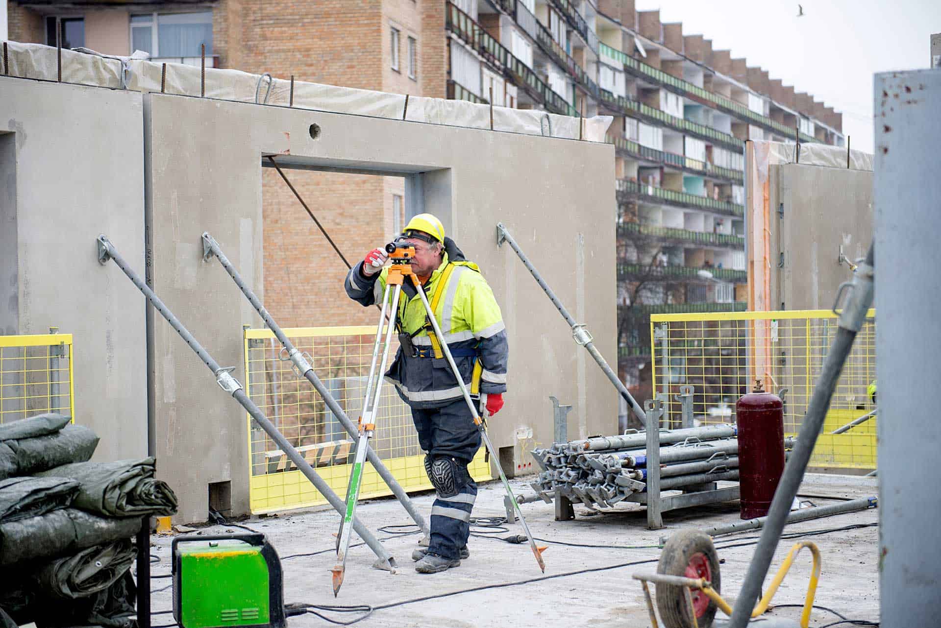 A construction worker, resembling a land surveyor in a yellow hard hat and high-visibility jacket, uses a leveling instrument on a bustling building site. Concrete walls and an array of construction materials border the urban backdrop of tall buildings.