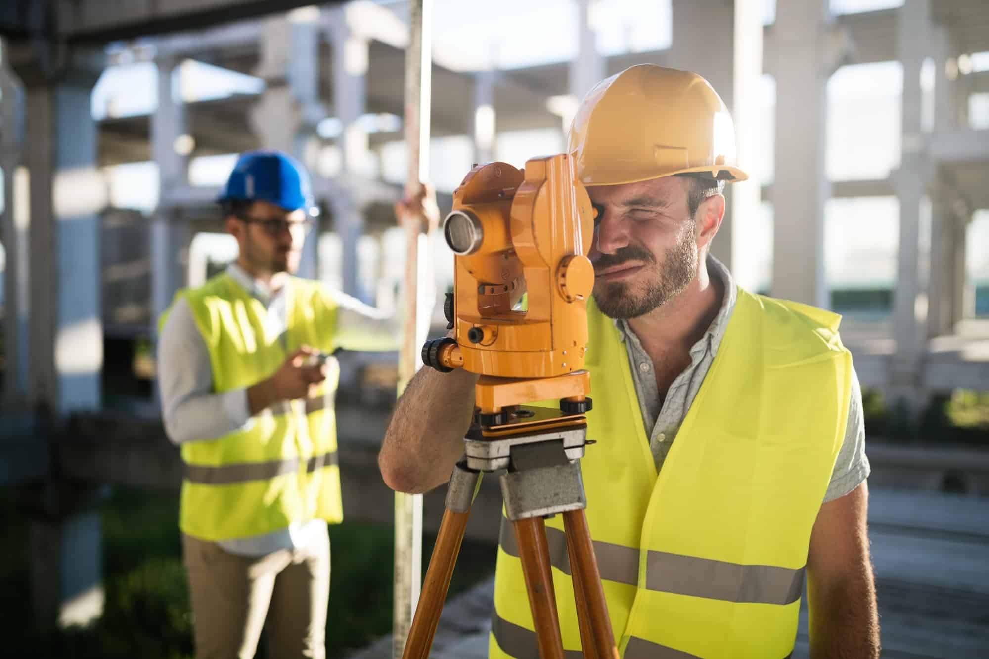 Two construction workers in high-visibility vests and helmets are busy on a construction site. One land surveyor uses a theodolite for precise measurements, while the other stands in the background holding a tablet. Concrete structures rise around them, forming the backdrop of their diligent work.