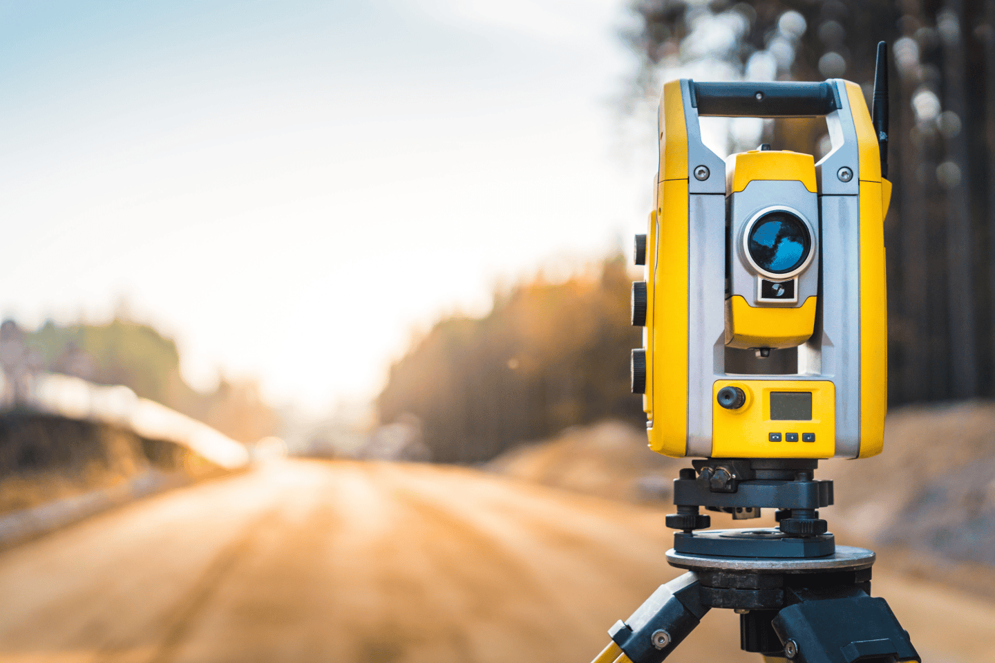 A yellow and gray total station is set up on a tripod, expertly positioned by a land surveyor on a construction site, with a blurred background of a dirt road and trees.