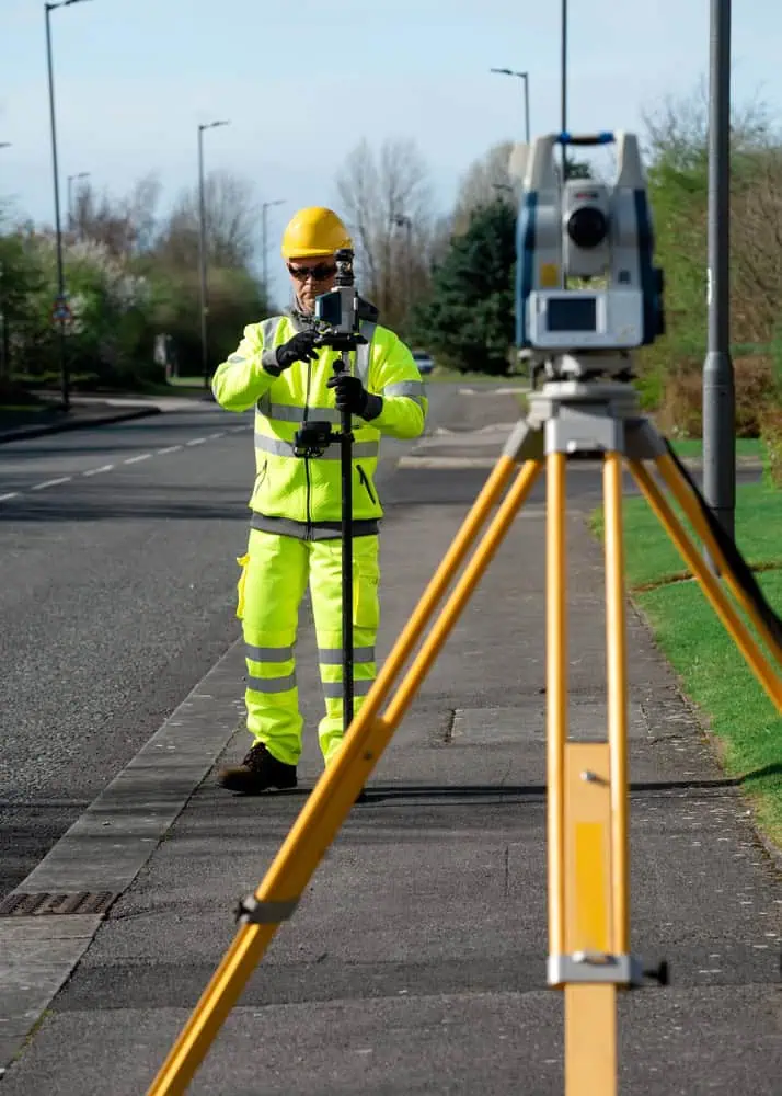 A land surveyor in a yellow safety suit and helmet stands on a sidewalk using surveying equipment. The tripod and total station are prominently positioned in the foreground, as the surveyor works alongside a tree-lined road.