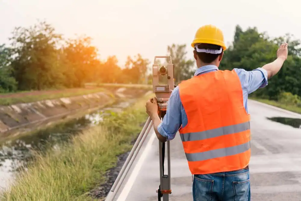 A land surveyor in a yellow hard hat and orange safety vest skillfully operates a tripod-mounted instrument on a rural road. The serene backdrop, featuring lush greenery and a tranquil canal, is beautifully illuminated by sunlight filtering through the trees.