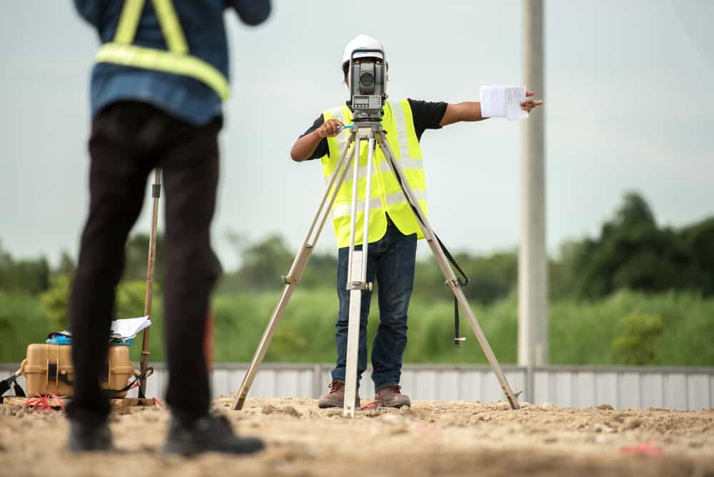 Two construction workers at a site: one, a land surveyor, operates a theodolite on a tripod, wearing a hard hat and high-visibility vest, pointing; the other stands in the foreground, blurred, dressed similarly. Trees and a fence frame the background.