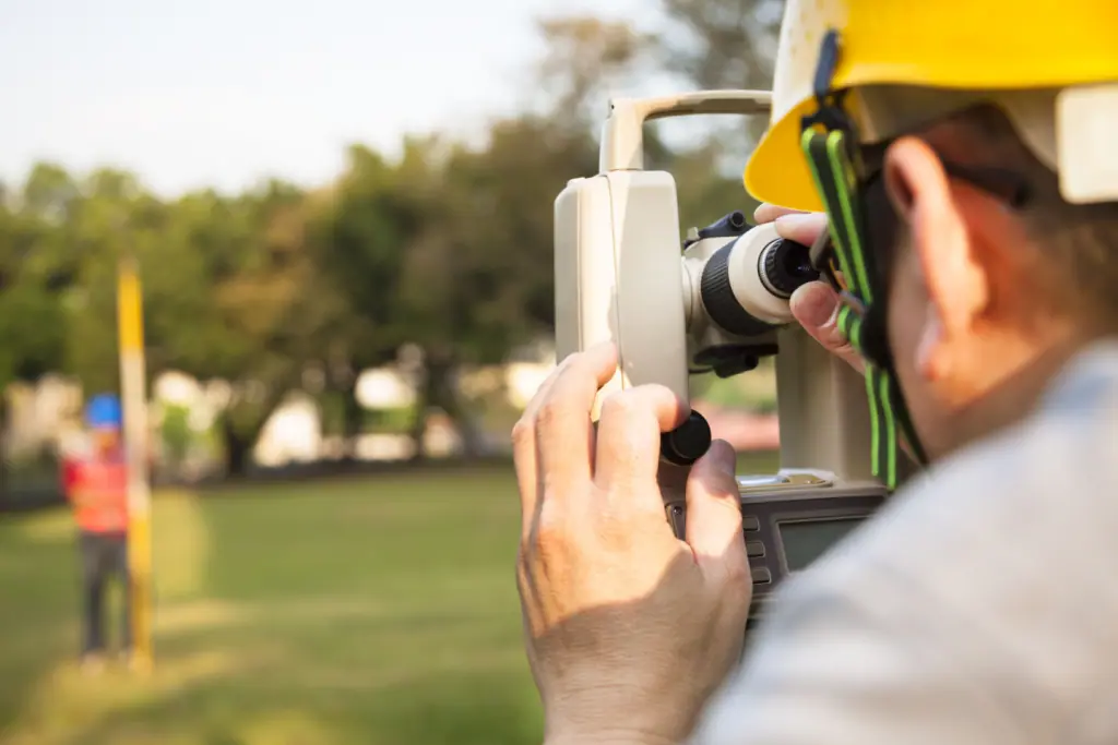 A land surveyor in a hard hat expertly uses a surveyor's instrument on a grassy field. In the background, another surveyor stands near a pole. The scene is set outdoors, framed by trees and a softly blurred landscape.