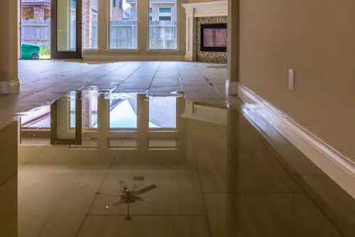 A living room with a large puddle of water on the tiled floor reflects the ceiling fan and sunlight streaming through the windows, as if a surveyor's precision was needed to truly capture its expanse. The room appears empty, with a fireplace visible in the background.