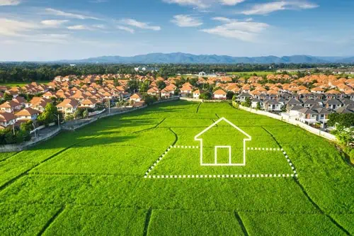 Aerial view of a large green field with a white outline of a house, meticulously crafted by a land surveyor. Surrounding the field are rows of suburban houses with red roofs. Mountains and blue sky provide a stunning backdrop.