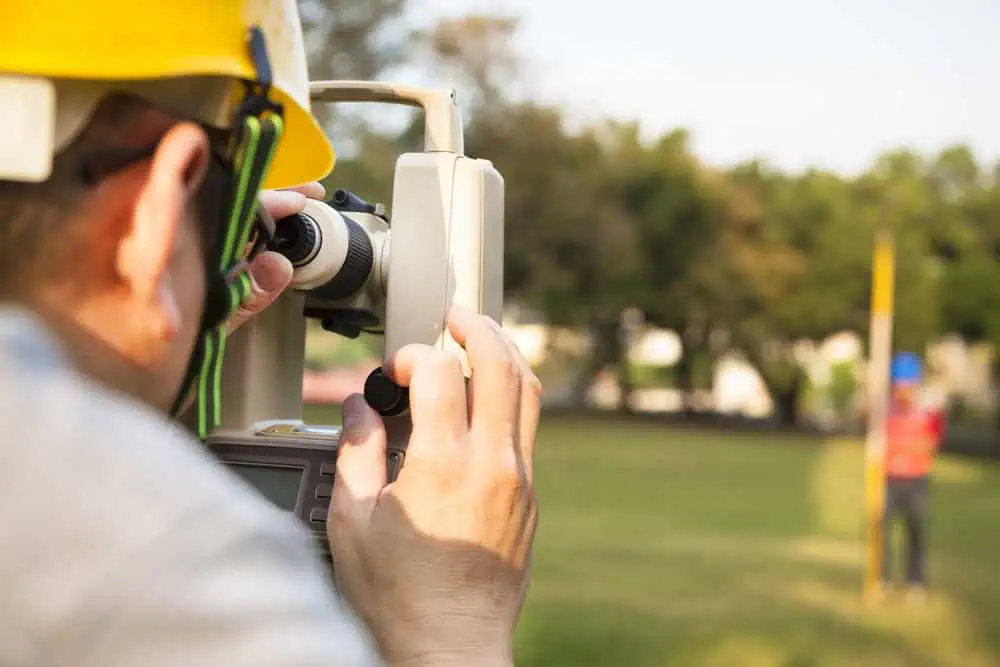 A surveyor wearing a yellow hard hat is using a theodolite to measure angles in an outdoor setting, with another person holding a measurement rod in the background on a grassy field. Trees and a clear sky are visible.