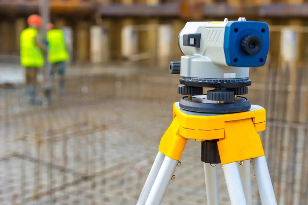 A theodolite on a tripod is set up at a construction site. In the blurred background, two workers in high-visibility vests and helmets are standing near rebar and concrete forms.