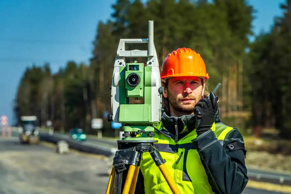 A construction worker wearing an orange hard hat and high-visibility vest operates a theodolite on a tripod. He is using a walkie-talkie. The background shows a partially constructed road lined with trees.
