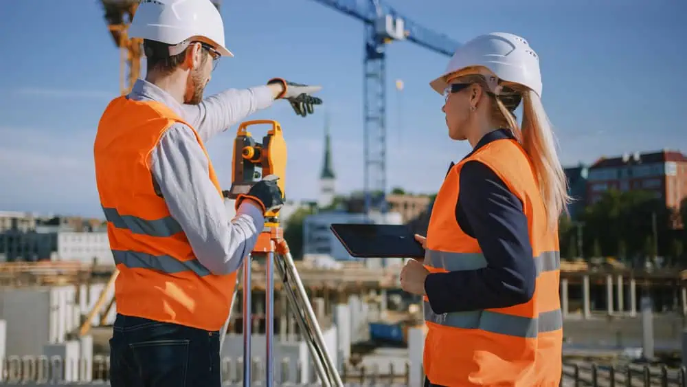 Two construction workers wearing white helmets and orange safety vests stand on a construction site. One holds a tablet, and the other points towards a crane in the background. A building structure is visible under a clear blue sky.