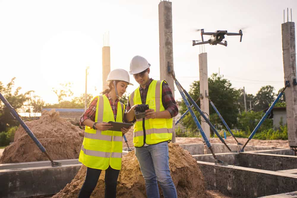 Two construction workers in safety vests and helmets examine a tablet and remote control at a building site. A drone flies in the background near concrete pillars under a clear sky.