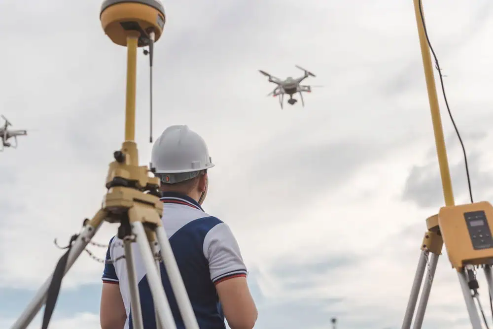 A person wearing a hard hat operates a drone in the sky using a remote control. Nearby are surveying equipment set up on tripods. The scene is set outdoors under a partly cloudy sky.