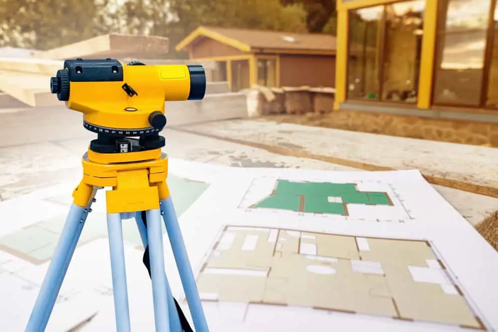 A yellow and black surveyor's level tripod stands on a wooden surface outdoors. Several architectural blueprints are laid out nearby. The background features wooden buildings with large windows.