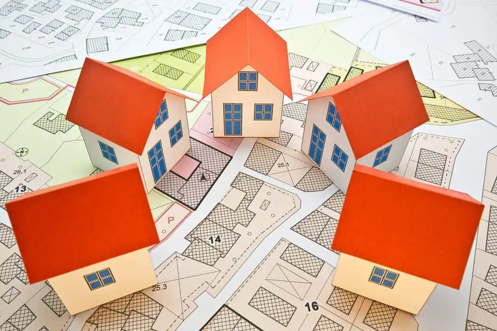 Five small model houses with red roofs and white walls are placed on architectural blueprints. The blueprints display various lot outlines and zoning patterns, suggesting urban planning or residential development concepts.