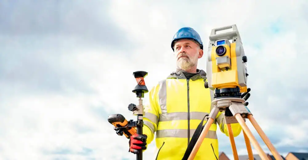 A man in a hard hat and high-visibility jacket operates surveying equipment on a tripod. He stands outdoors under a cloudy sky, holding a surveying tool in one hand and looking into the distance.