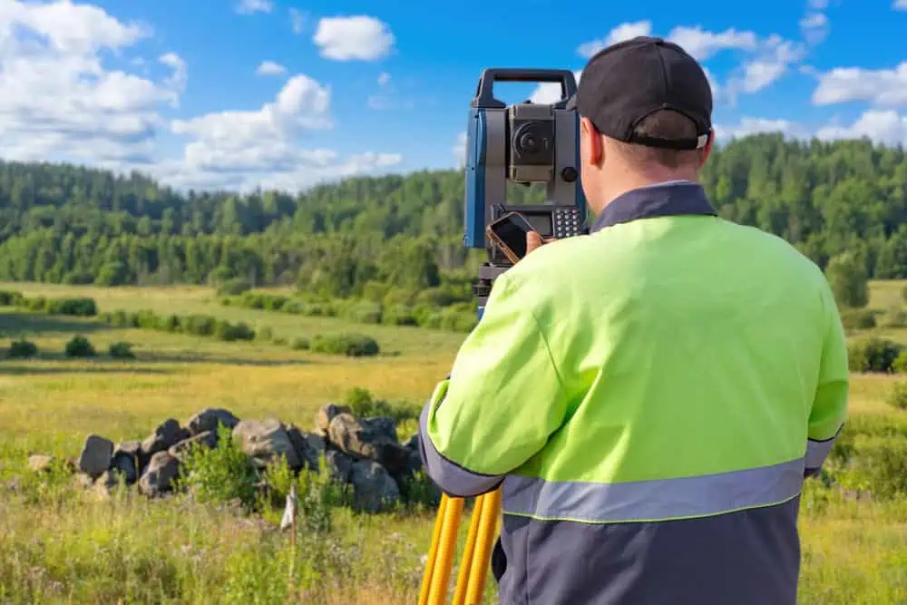 A surveyor wearing a green jacket and black cap uses a theodolite on a tripod in a scenic countryside. The background features a stone pile, lush green trees, and a bright blue sky with clouds.