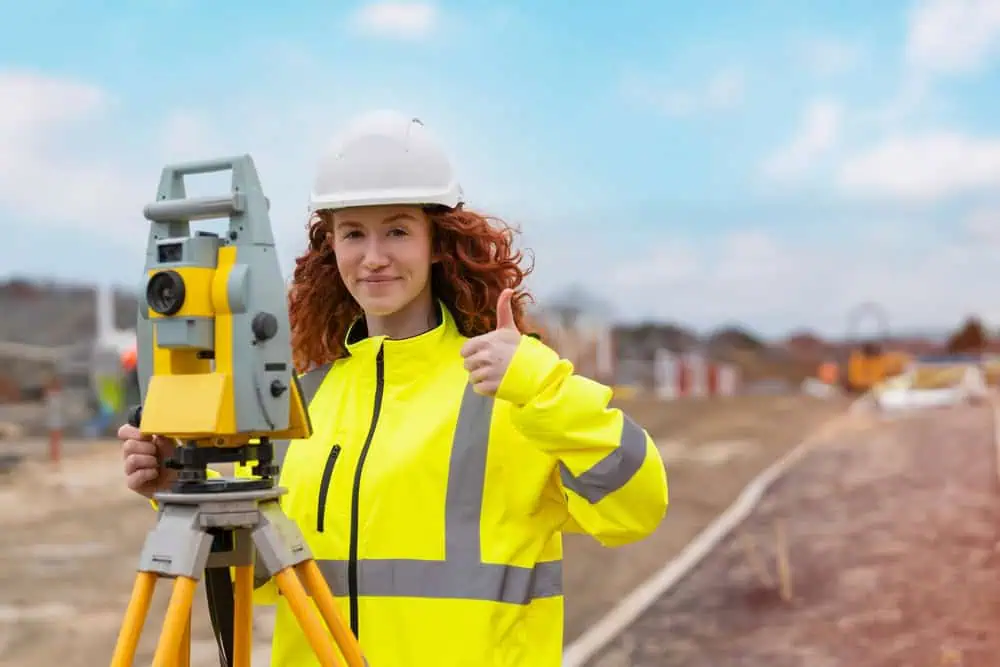A smiling woman in a bright yellow safety jacket and white hard hat gives a thumbs up while standing next to survey equipment on a construction site. The background shows a blurred view of the site under a blue sky.