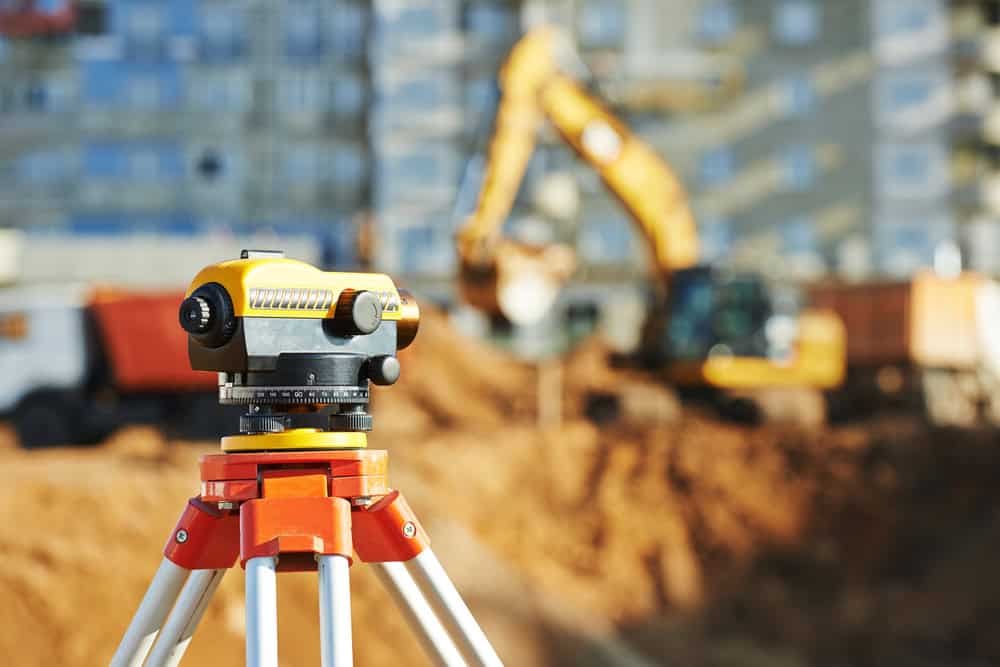 A surveying instrument on a tripod is in focus in the foreground, with a construction site featuring an excavator and buildings blurred in the background. The scene suggests preparation or measurement for construction work.