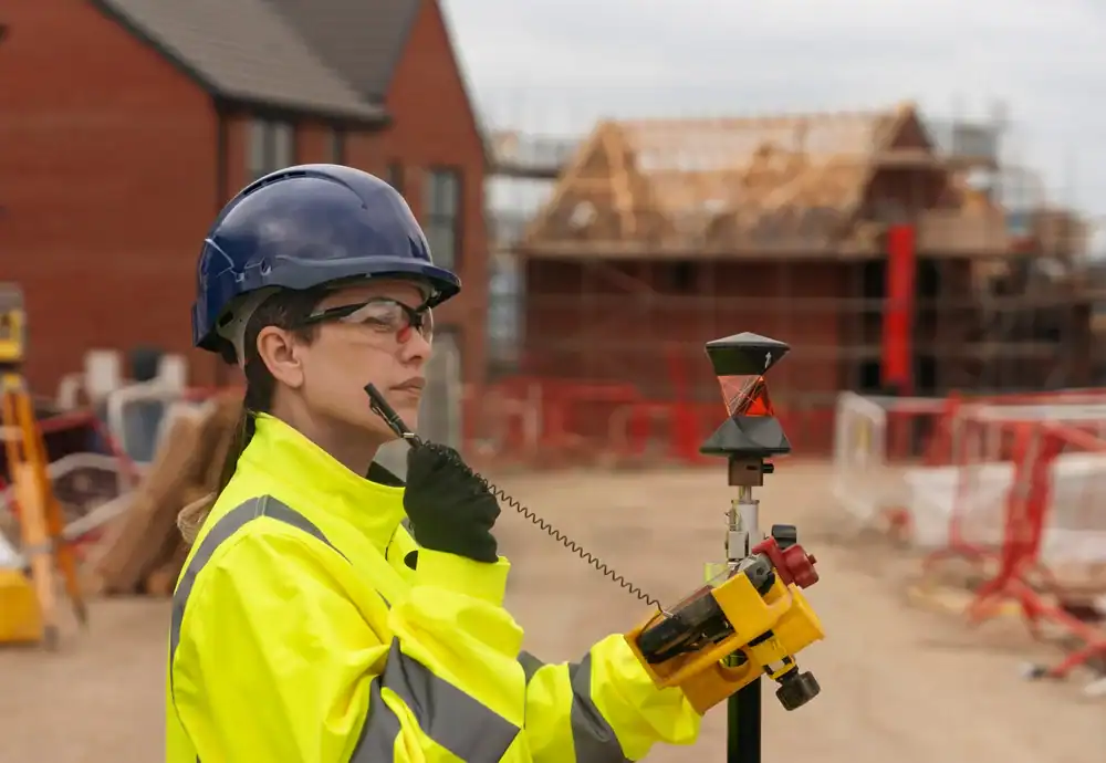A construction worker in a high-visibility jacket and safety gear uses a walkie-talkie while operating surveying equipment at a building site with partially constructed houses in the background.
