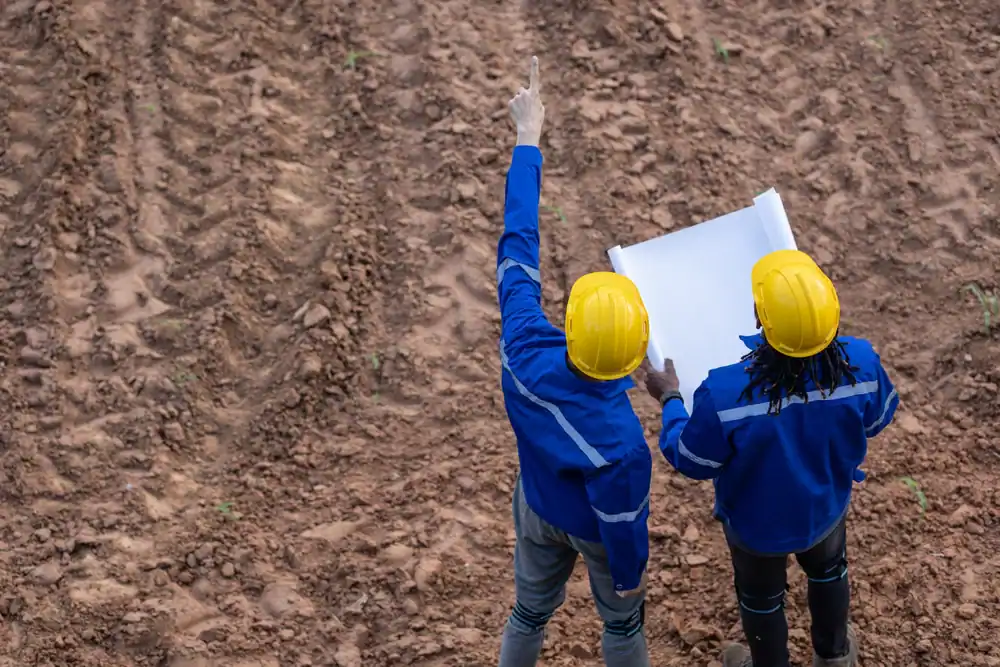 Two construction workers in blue uniforms and yellow helmets stand on a dirt field. One holds blueprints while the other points ahead, discussing plans or directions on the construction site.