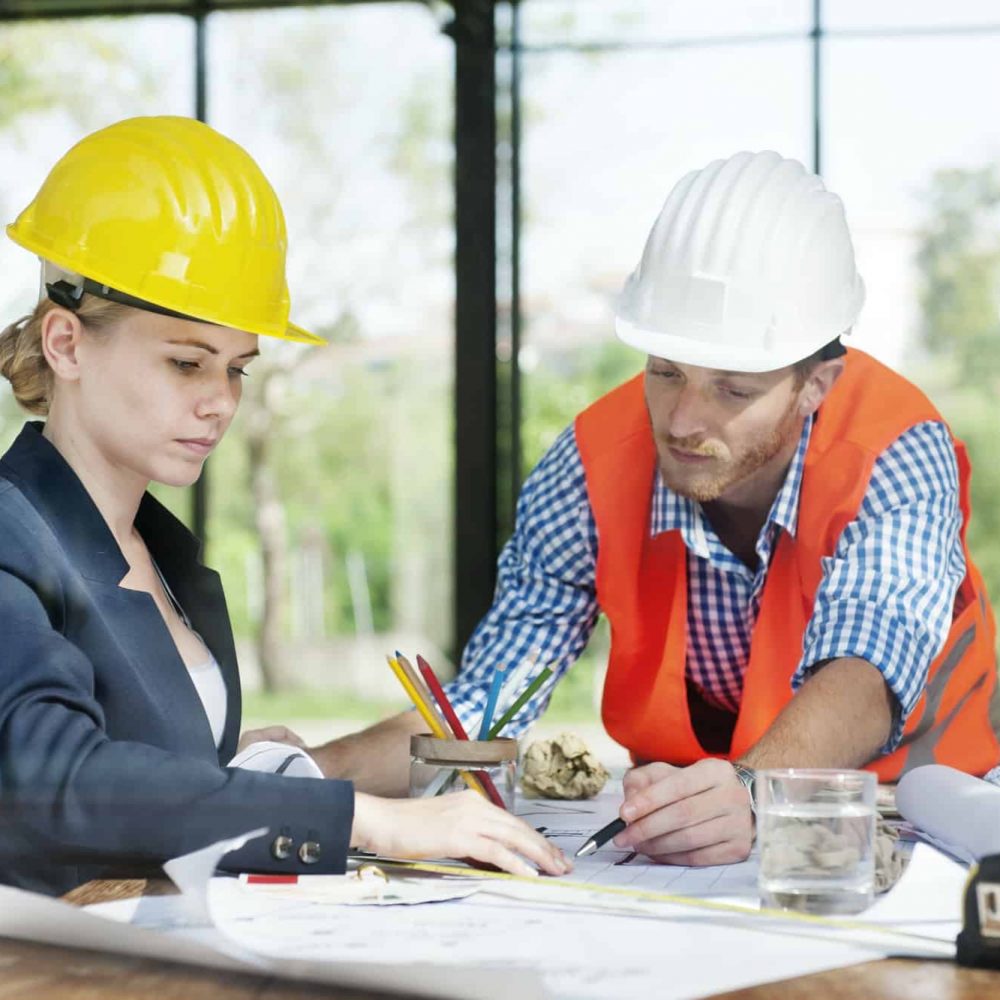 A man and a woman in hard hats, possibly land surveyors, are working at a table covered with architectural plans and pencils. The man, dressed in an orange vest, points at the plans as sunlight filters through large windows showcasing greenery outside.