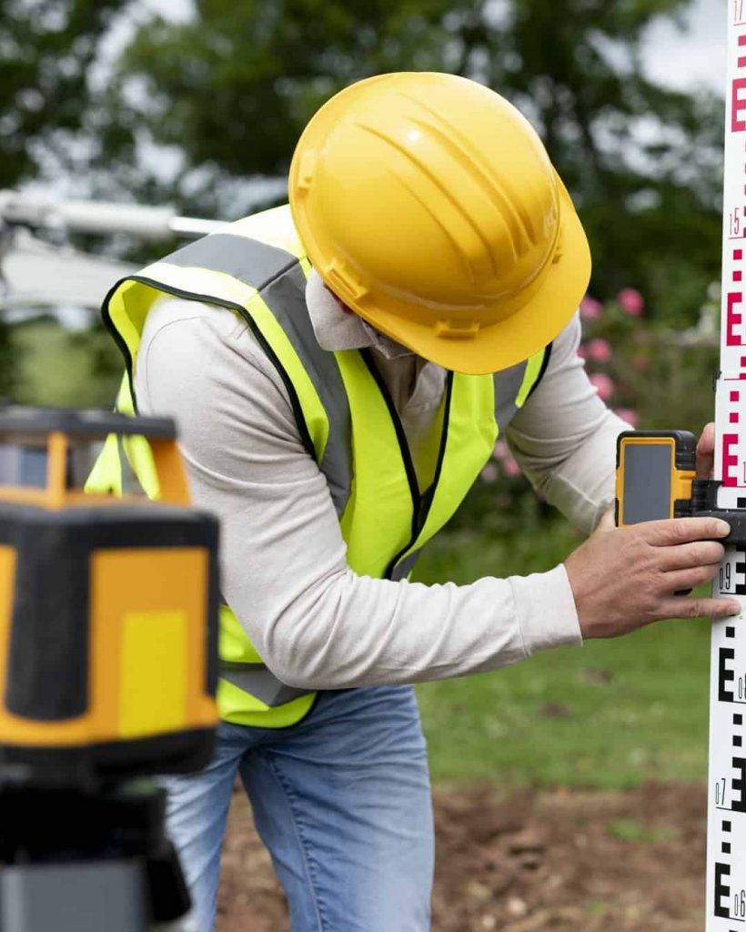 A construction worker, resembling a dedicated land surveyor, in a yellow hard hat and reflective vest adjusts a measuring instrument on a leveling rod. A laser level is positioned in the foreground, with trees and construction equipment visible in the background.