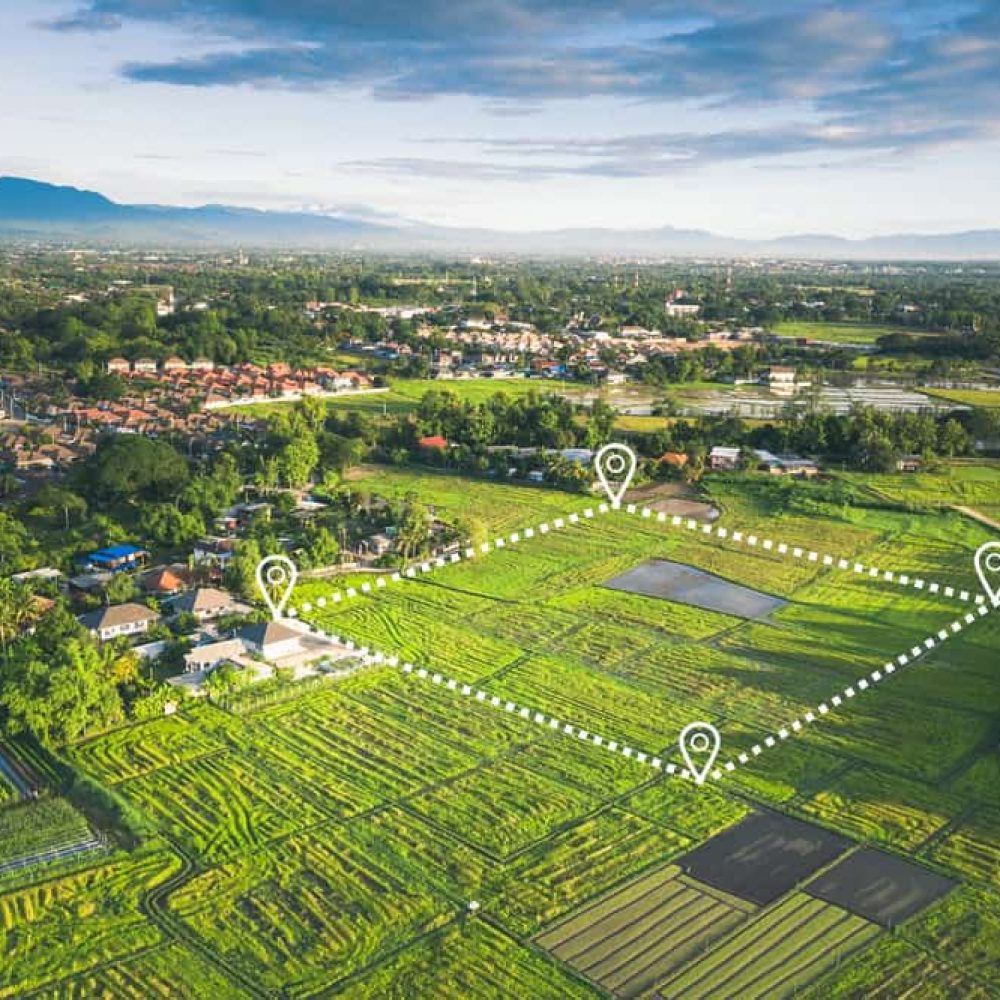 Aerial view of a lush green agricultural landscape with modern technology graphics highlighting specific plots, reminiscent of a surveyor's precision. The area is surrounded by residential buildings and mountains visible in the background under a blue sky with scattered clouds.