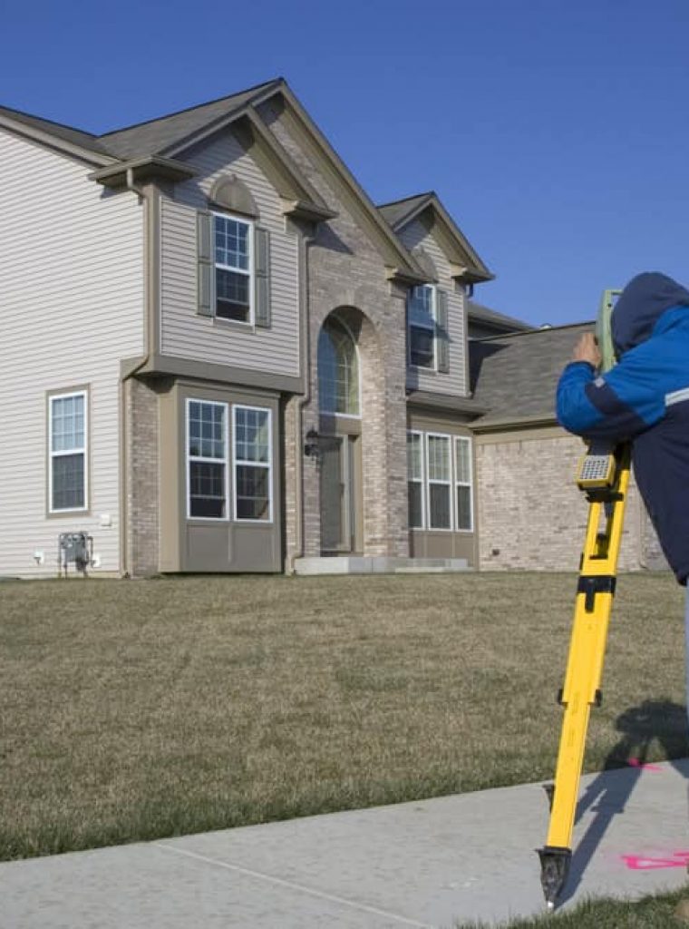 A land surveyor in a blue jacket and jeans uses a yellow tripod and equipment in front of a large suburban house with gray siding and brick accents. With manicured lawns under a clear blue sky, this two-story home epitomizes modern elegance while the surveyor carefully measures boundaries.
